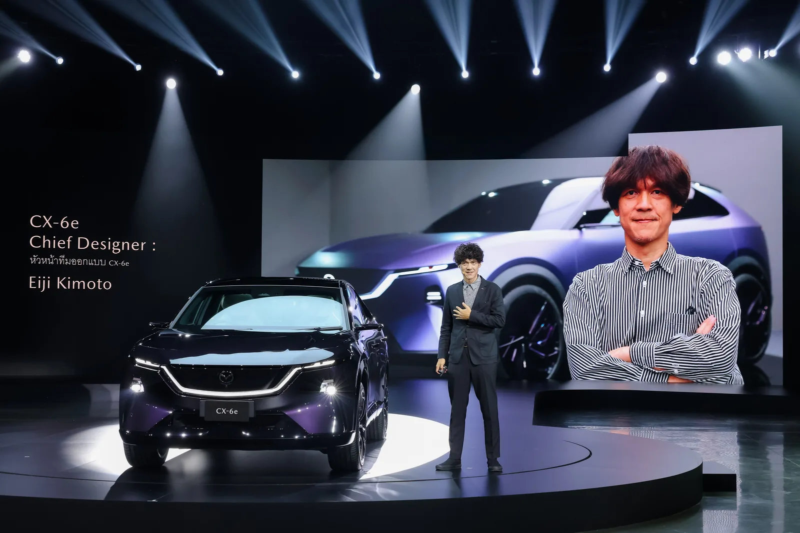 Eiji Kimoto หัวหน้าทีมออกแบบ Mazda CX-6e ยืนบนเวทีงานเปิดตัวข้างตัวรถ