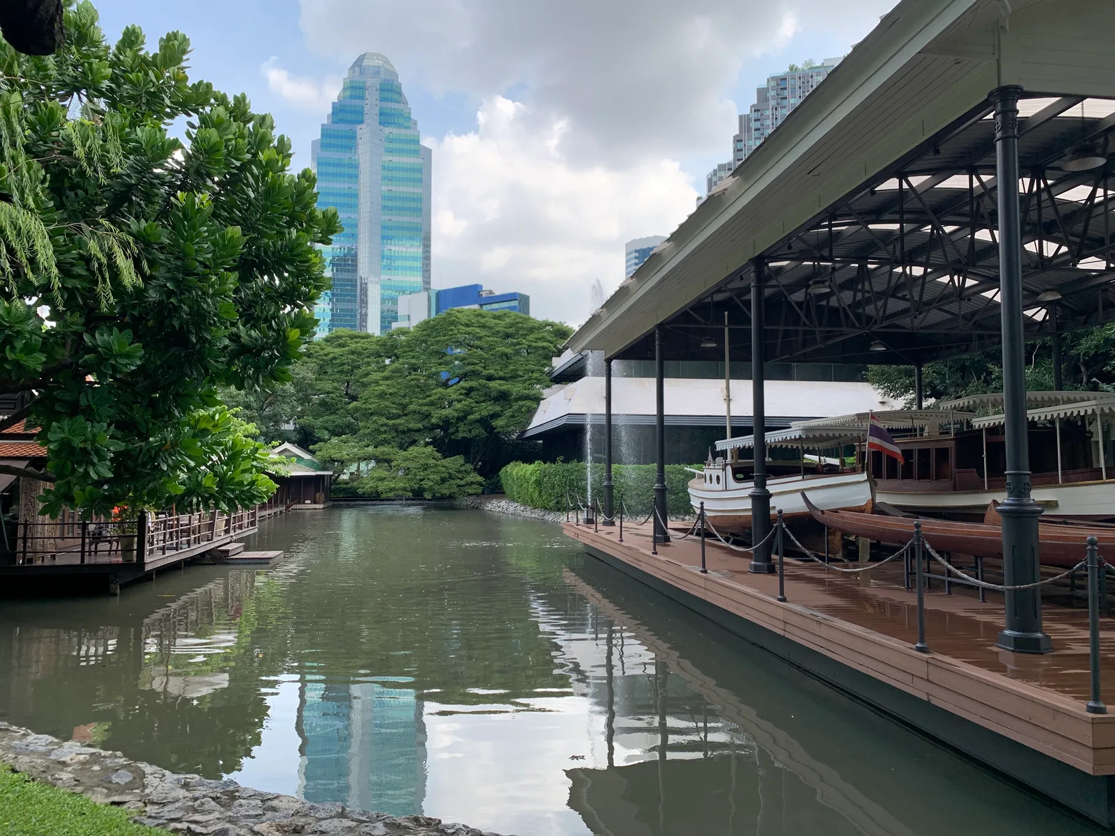 สระน้ำยาวในบ้านปาร์คนายเลิศ ข้างโรงเรือหลังคาโครงเหล็กที่มีเรือเก่าจอดอยู่ ด้านหลังเป็นตึกสูง