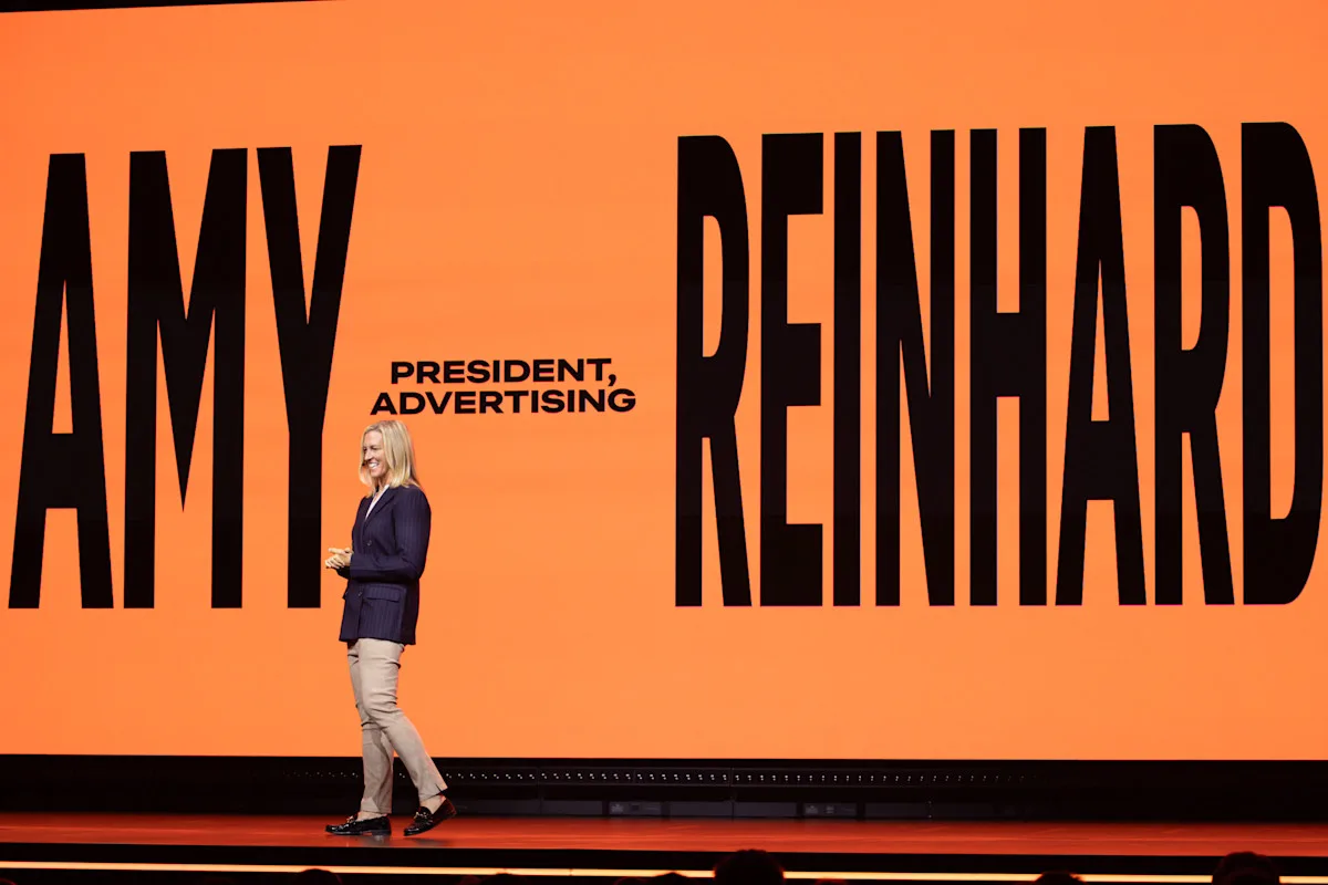 Amy Reinhard ประธานฝ่ายโฆษณาของ Netflix ยืนพูดบนเวทีหน้าจอสีส้มที่เขียนชื่อและตำแหน่ง President, Advertising ในงาน UK Upfront ของ Netflix