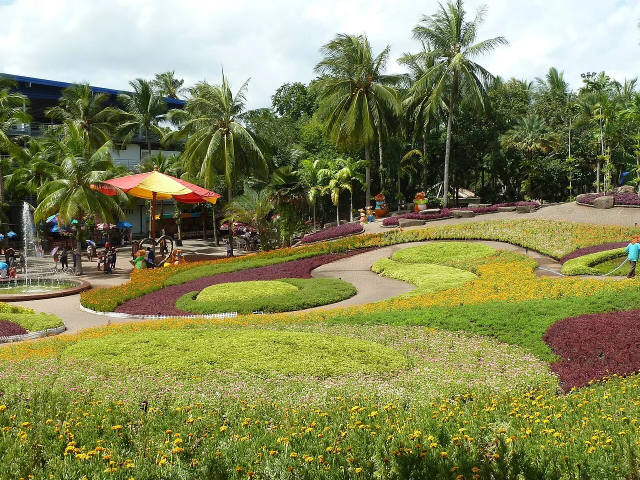 ผืนดอกไม้หลากสีจัดเป็นลวดลายโค้งในสวนนงนุช มีต้นมะพร้าวและร่มสีสดด้านหลัง