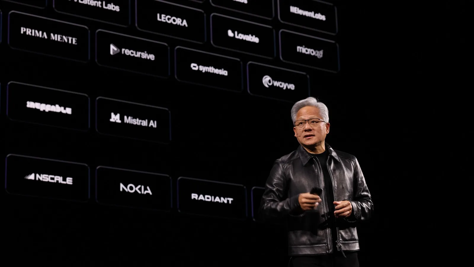 Jensen Huang ซีอีโอของ NVIDIA ในเสื้อแจ็กเก็ตหนังสีดำ ยืนพูดบนเวทีหน้าจอที่แสดงโลโก้บริษัทพันธมิตร เช่น Mistral AI, Nokia และ Wayve