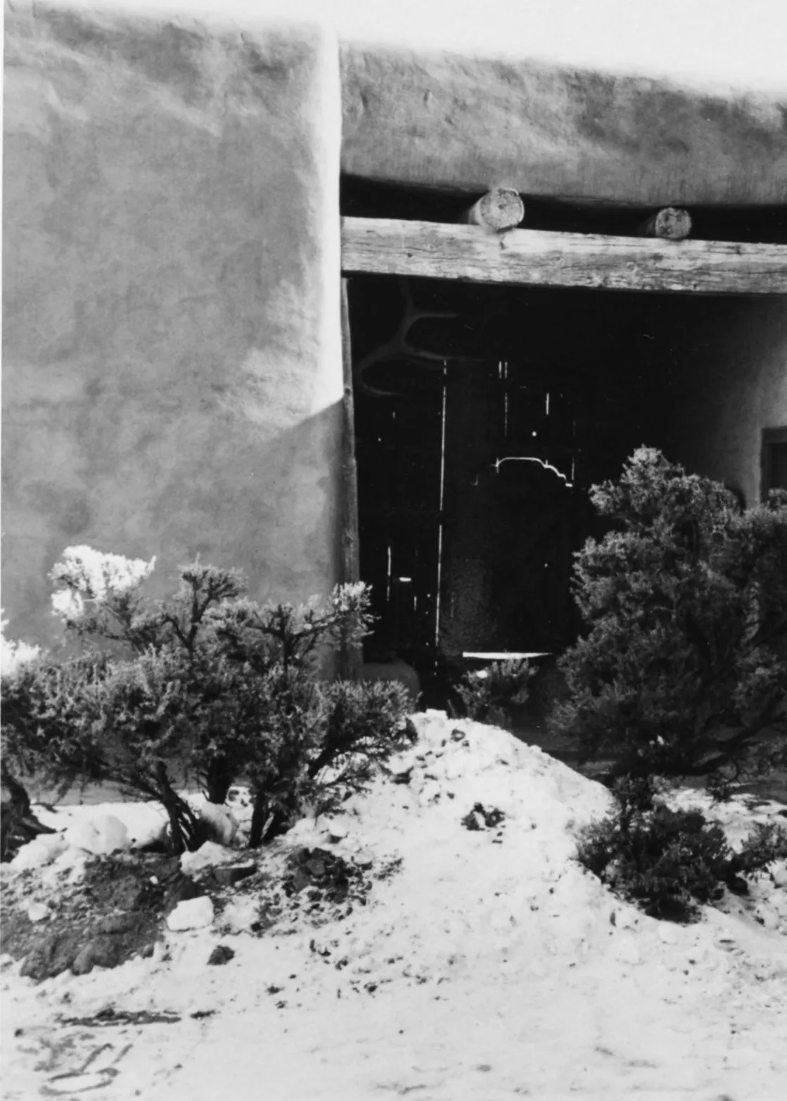 ลานกลางของบ้าน Georgia O'Keeffe ที่ Abiquiú มองไปทางโถงทางเข้าหลัก ผนังดินอะโดบี คานไม้ และพุ่มไม้บนพื้นที่มีหิมะ