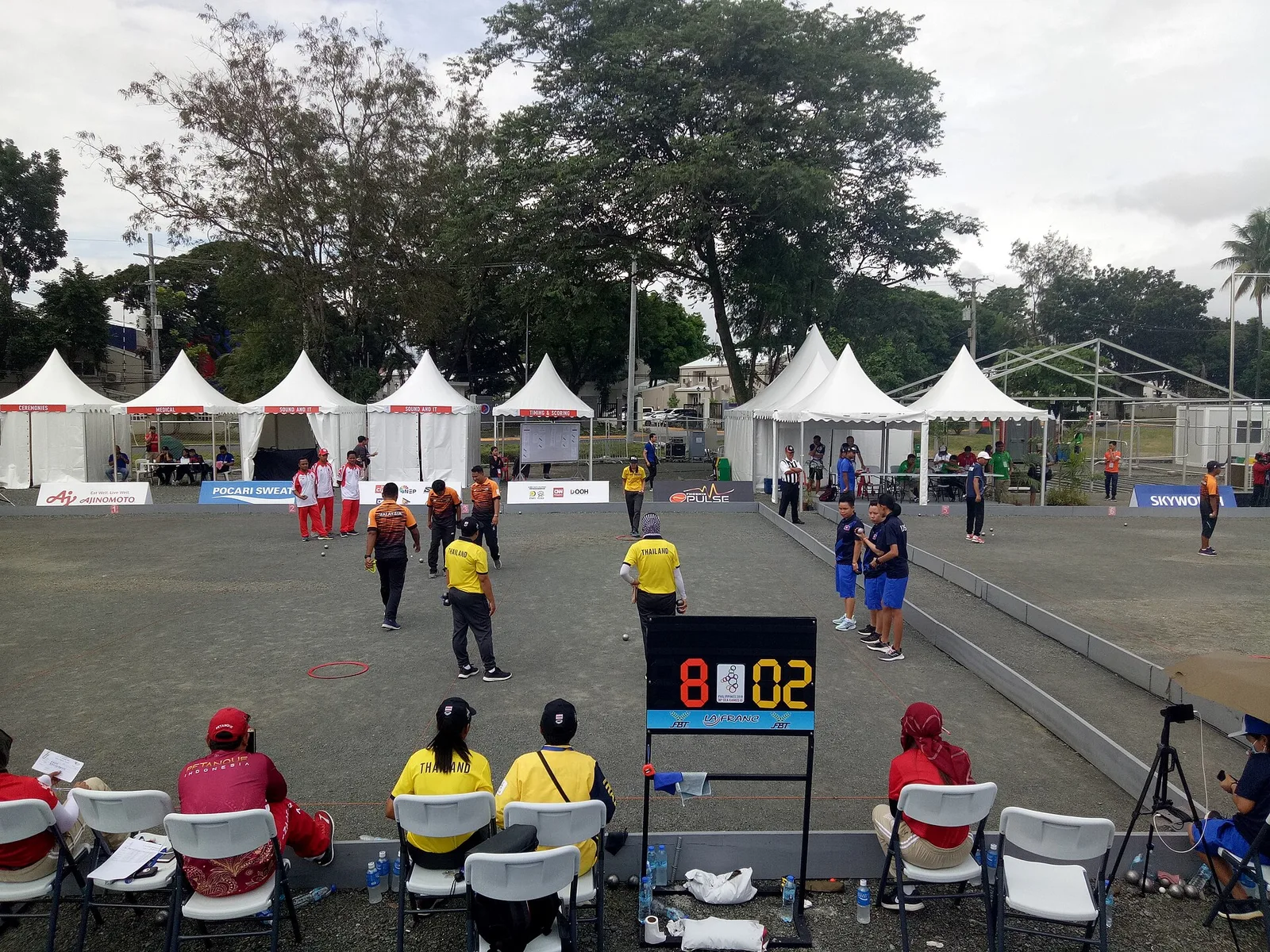 ลานเปตองในซีเกมส์ 2019 ที่ Clark Freeport ฟิลิปปินส์ นักเปตองหญิงไทยในเสื้อโปโลสีเหลืองกางเกงดำแข่งประเภททีม 3 คนกับลาวในเสื้อสีกรมท่า มีวงโยนสีแดงบนพื้นกรวดและป้ายคะแนน 8-02