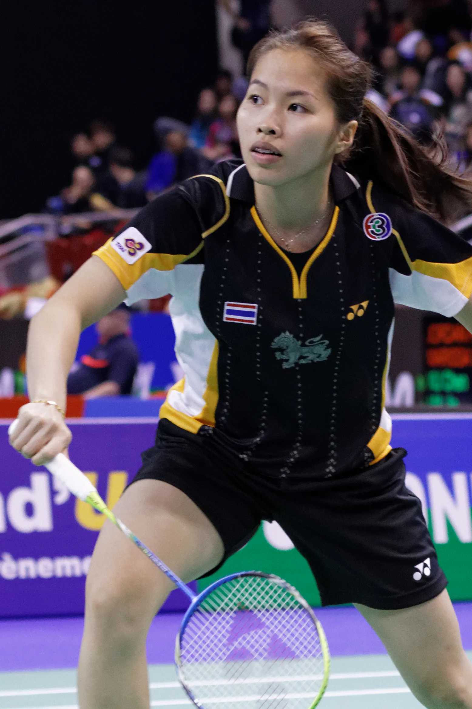 รัชนก อินทนนท์ สวมเสื้อแข่งทีมชาติไทยสีดำเหลืองติดธงชาติและกางเกงขาสั้น ถือแร็กเกตแบดมินตันในท่าเตรียมรับลูก ในรอบก่อนรองชนะเลิศหญิงเดี่ยว French Open ปี 2013