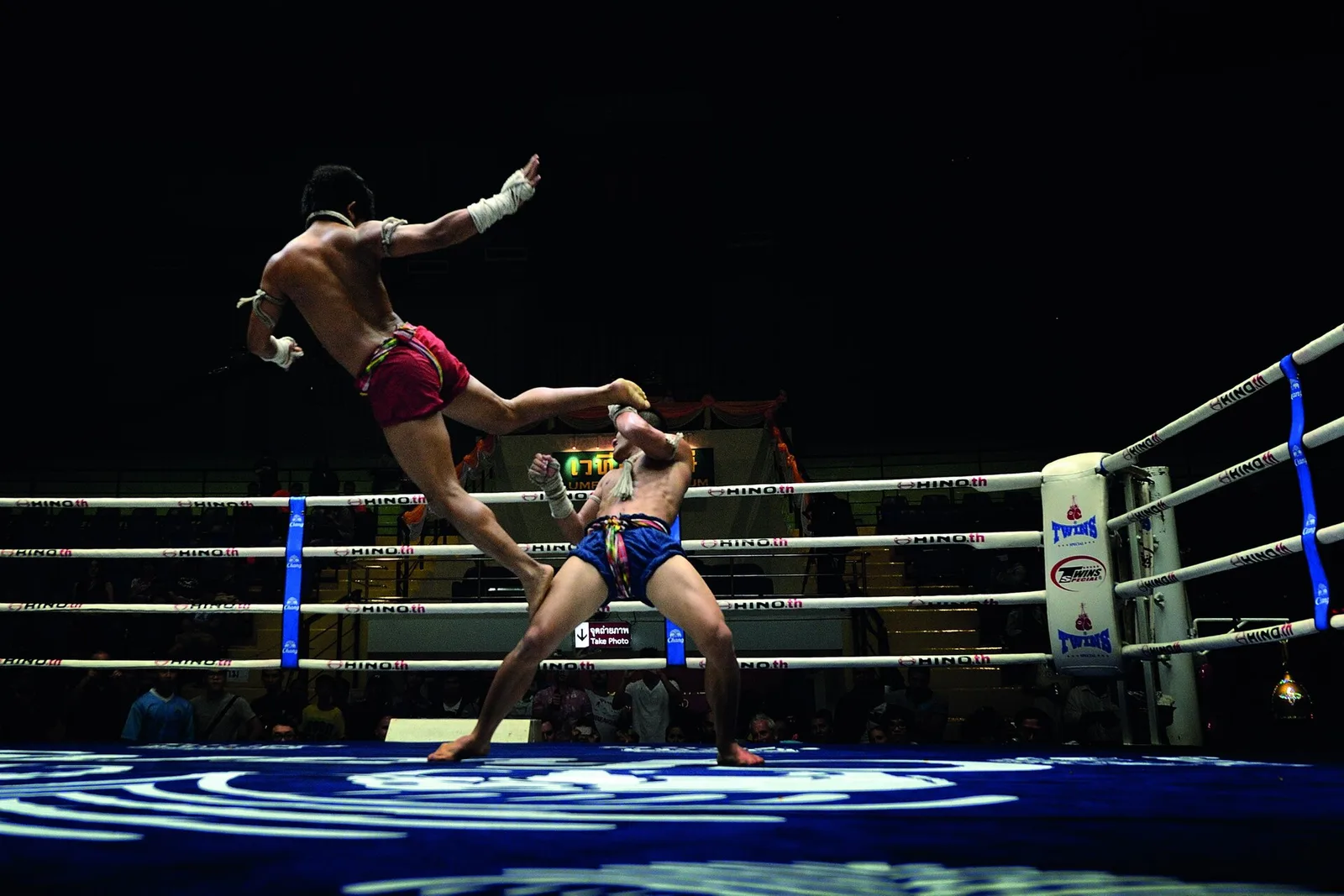 นักมวยไทยสวมกางเกงมวยสีแดงกระโดดเตะคู่ชกกางเกงสีน้ำเงินบนเวทีสนามมวยเวทีลุมพินี ภาพถ่ายเมื่อ 16 มกราคม 2015