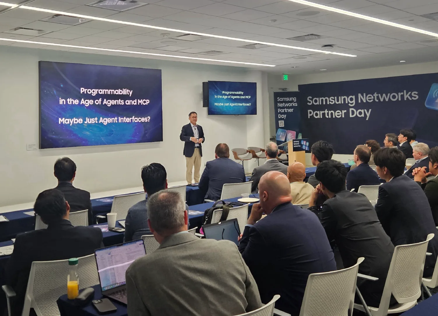 ผู้บรรยายยืนหน้าจอในห้องประชุมงาน Samsung Networks Partner Day โดยมีผู้เข้าร่วมนั่งฟังและป้ายชื่องานอยู่ด้านหลัง