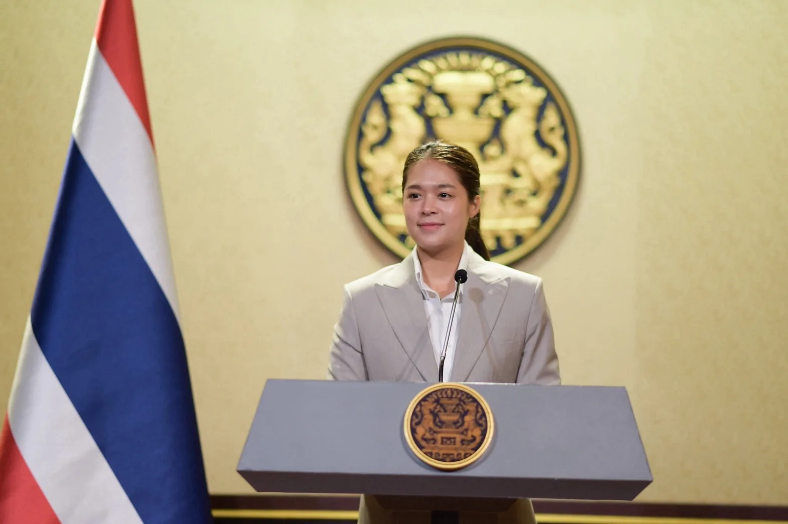 รองโฆษกประจำสำนักนายกรัฐมนตรีผู้แถลงเรื่องเที่ยวทั่วไทยไปกับทัวร์ ยืนที่โพเดียมหน้าตราสัญลักษณ์ของทำเนียบรัฐบาล ข้างธงชาติไทย