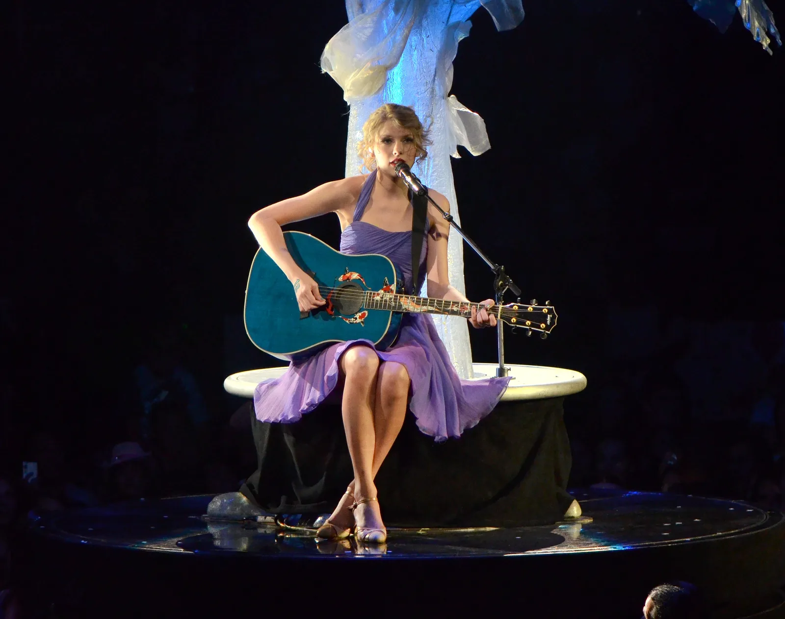 Taylor Swift นั่งเล่นกีตาร์ในชุดสีม่วงบนเวที Speak Now World Tour เดือนกรกฎาคม 2011