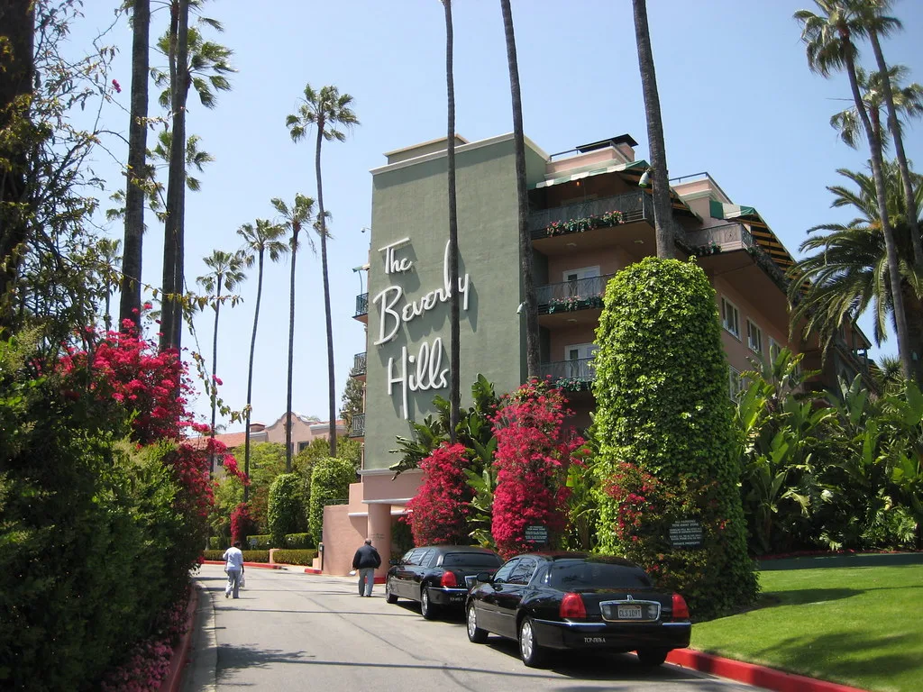 โรงแรม The Beverly Hills Hotel อาคารสีชมพูแซมเขียวมีป้ายชื่อโรงแรม แนวต้นปาล์มและดอกเฟื่องฟ้าริมทางรถเข้า ภาพวันที่ 27 เมษายน 2008