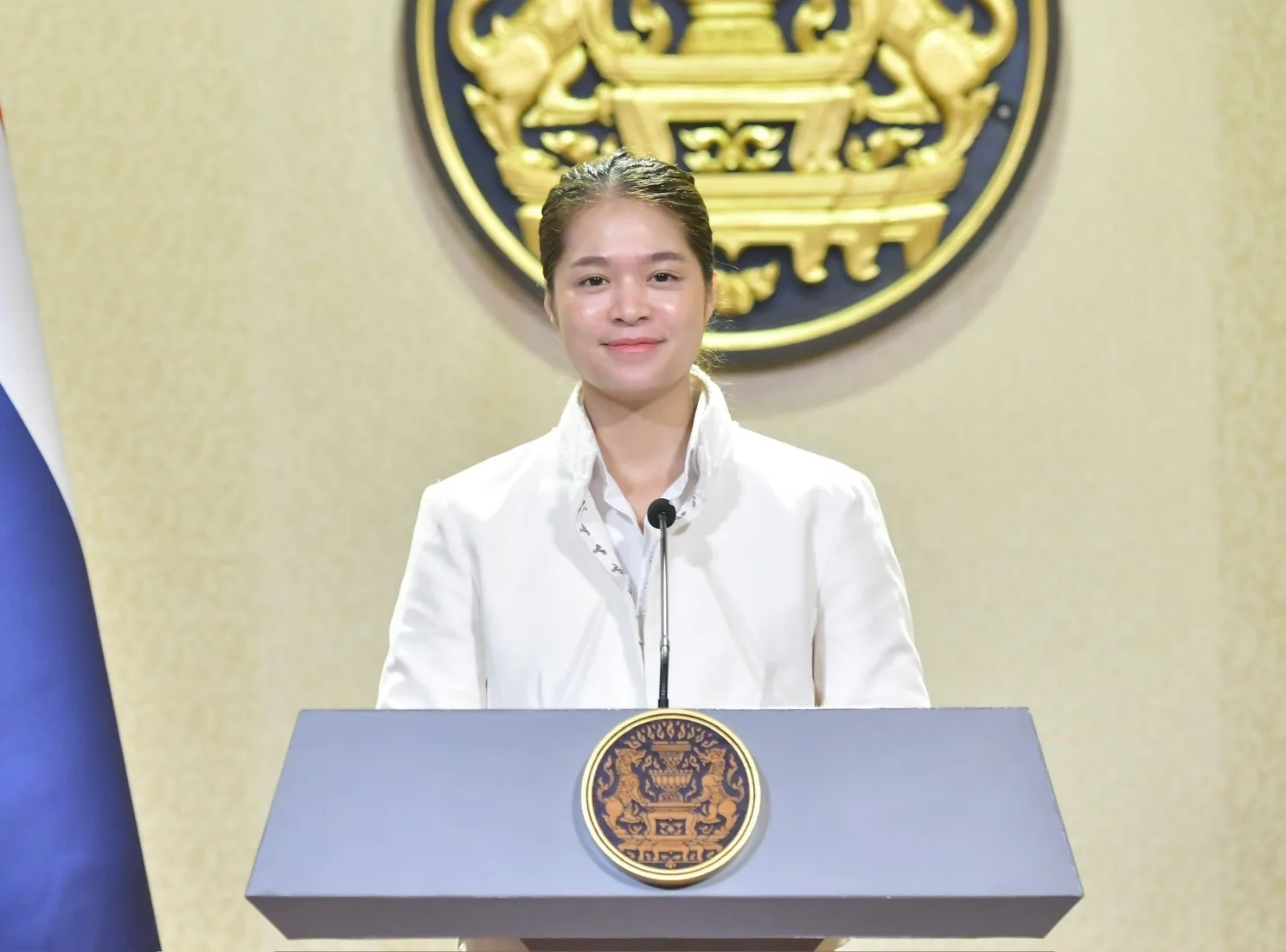 รองโฆษกประจำสำนักนายกรัฐมนตรียืนหลังโพเดียมที่มีตราสัญลักษณ์รัฐบาล ภาพประกอบข่าวรัฐบาลขยายโครงการแฟรนไชส์ไทยช่วยไทย 10,000 บาท ถึงเดือนธันวาคม 2570