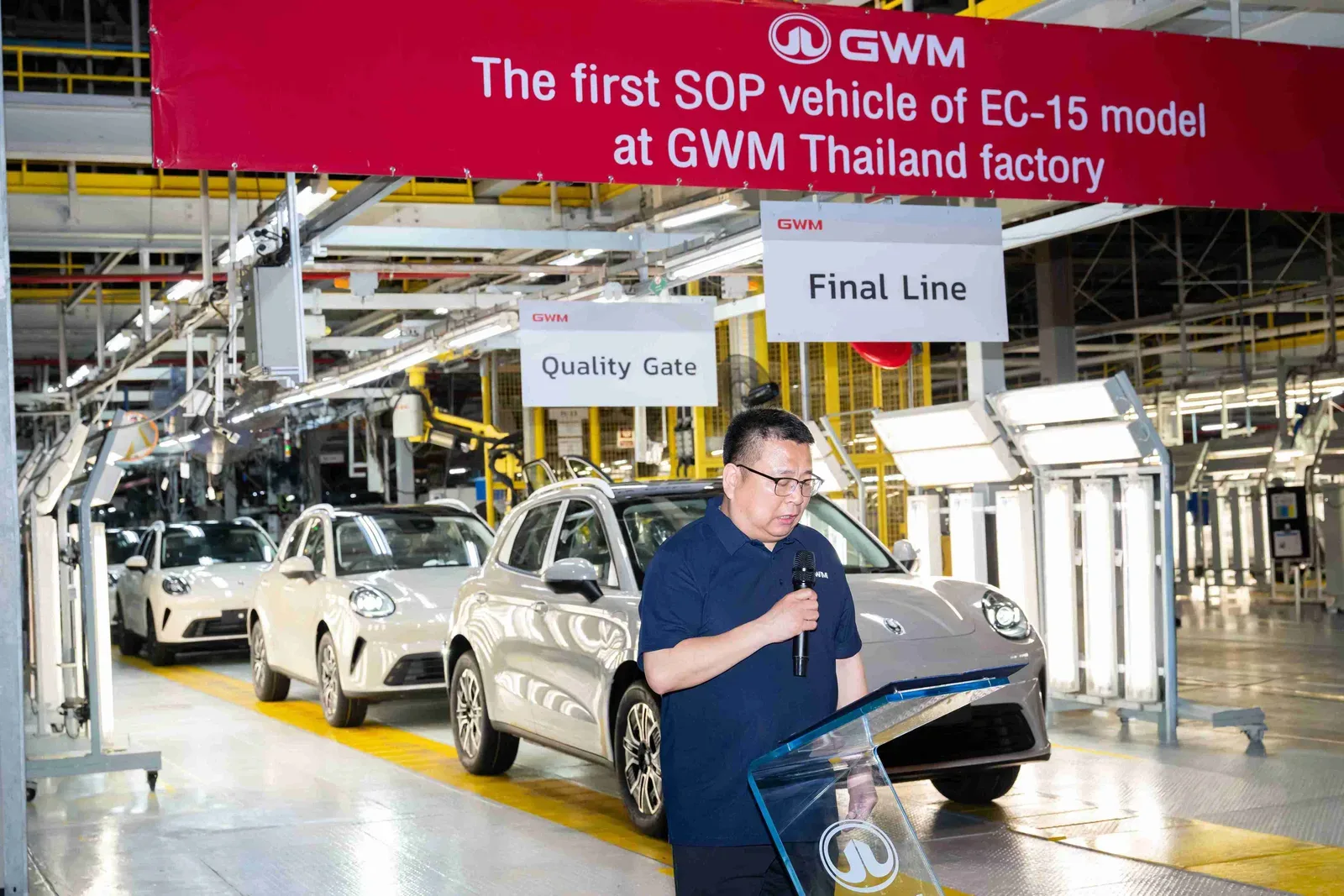 ผู้บริหาร GWM กล่าวบนแท่นหน้ารถ ORA 5 สีเงินและสีขาวที่สายประกอบ Final Line ใต้ป้าย The first SOP vehicle of EC-15 model at GWM Thailand factory