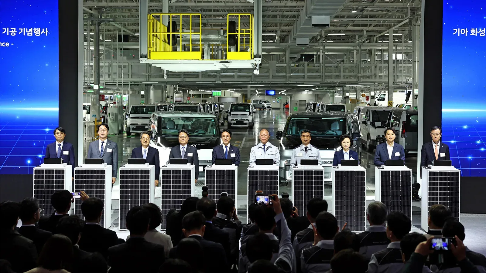 พิธีในโรงงาน Hwaseong EVO Plant ของ Kia ผู้บริหารยืนเรียงแถว จอด้านข้างมีข้อความภาษาเกาหลี และมีรถตู้ PV5 หลายคันจอดอยู่ด้านหลัง