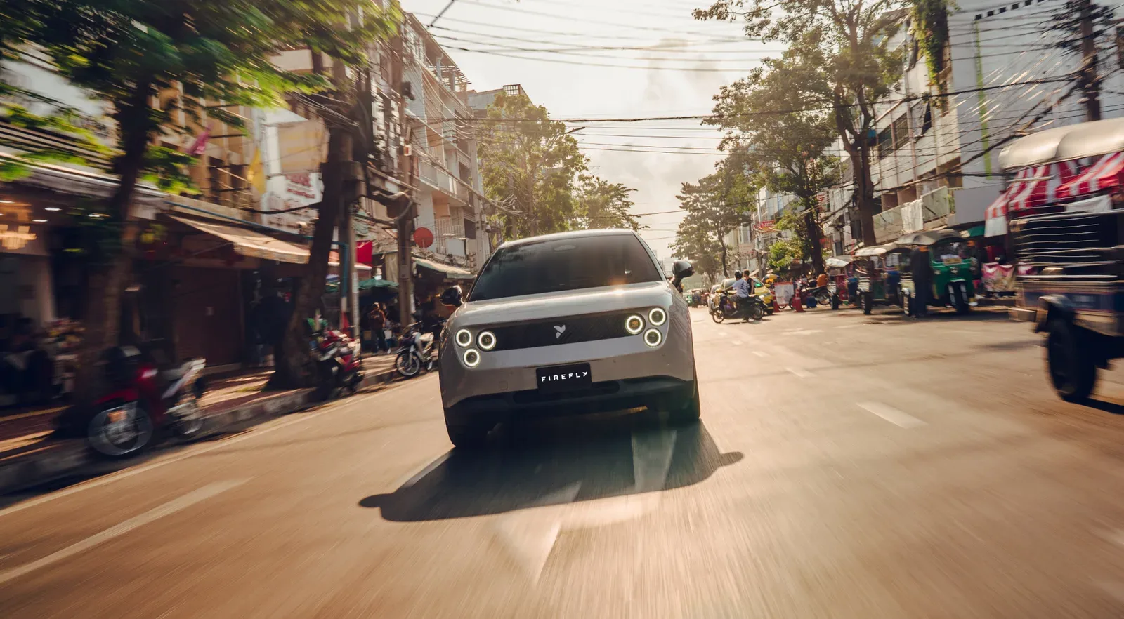 NIO firefly สีเงินวิ่งบนถนนในเมืองที่มีรถตุ๊กตุ๊กและร้านค้าสองข้างทาง ภาพจากเว็บไซต์ firefly ประเทศไทย