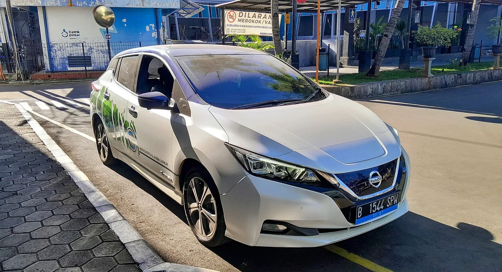 Nissan LEAF รุ่นที่ 2 (ZE1) สีขาวติดสติกเกอร์มหาวิทยาลัยบูดีลูฮูร์ จอดริมถนนภายในมหาวิทยาลัย มีนาคม 2024