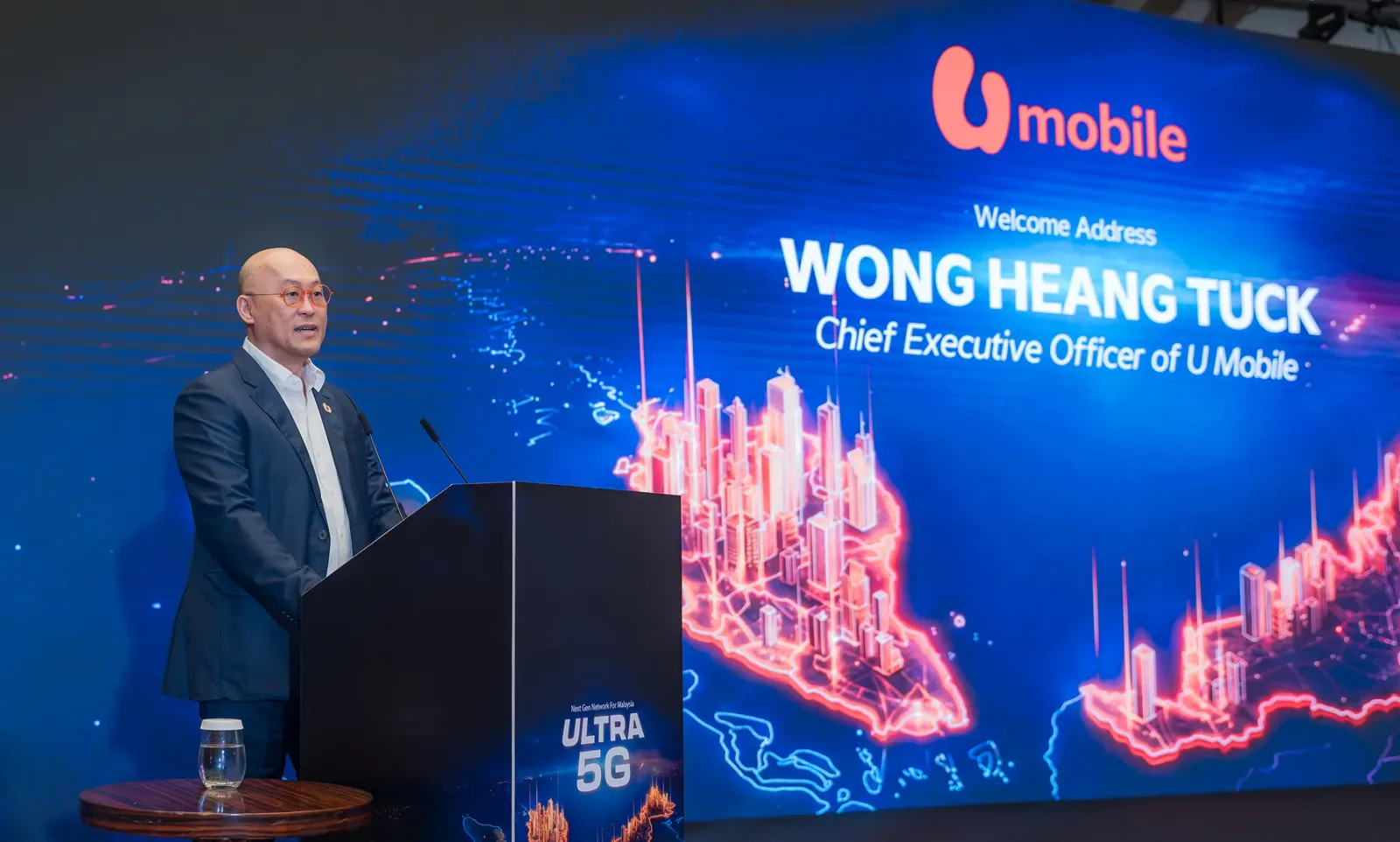 Wong Heang Tuck ซีอีโอของ U Mobile กล่าวต้อนรับในงานประกาศความครอบคลุม 5G 90% บนเวทีที่มีโลโก้ ULTRA 5G