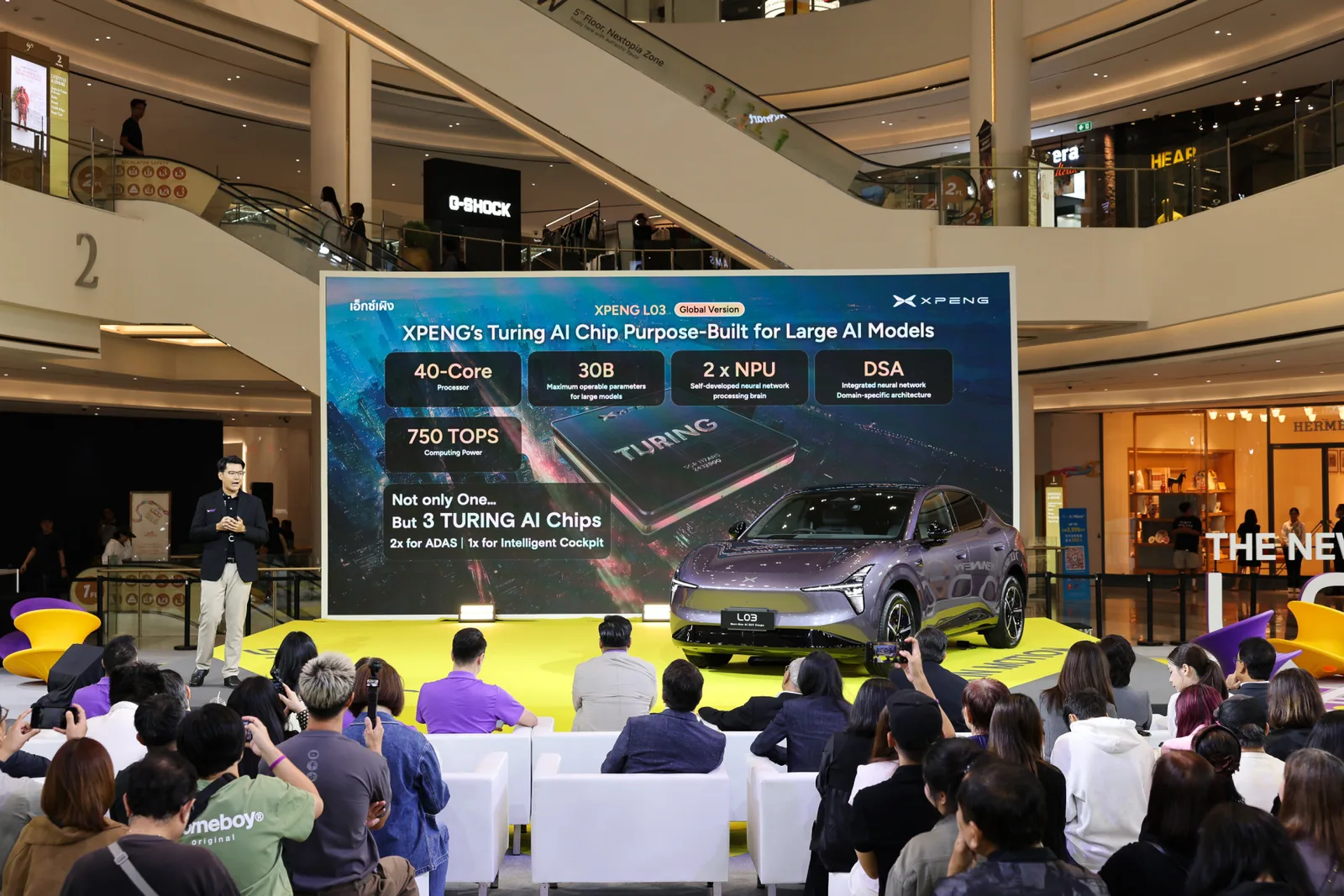 จอนำเสนอชิป Turing AI ของ XPENG บนเวทีงานเปิดตัว XPENG L03 มีผู้ชมนั่งฟังและรถจอดอยู่หน้าจอ