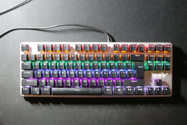 คีย์บอร์ดเมคานิคอลแบบไม่มีปุ่มตัวเลขสีดำ มีไฟ RGB หลายสีใต้ปุ่ม วางบนโต๊ะสีเข้ม