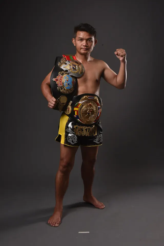 นักมวยไทยยืนในสตูดิโอพื้นหลังเทาเข้ม สวมกางเกงมวยสีดำเหลือง คาดเข็มขัดแชมป์รอบเอวและพาดเข็มขัดอีกเส้นที่ไหล่