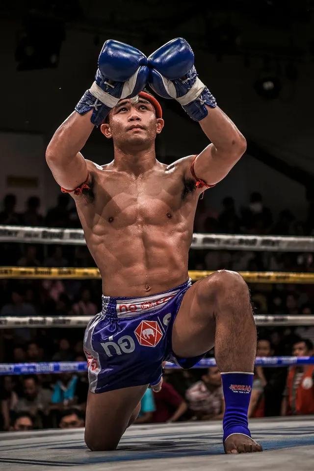 นักมวยไทยคุกเข่าบนเวที ยกนวมสีน้ำเงินเหนือศีรษะ สวมกางเกงมวยสีน้ำเงินขาว ประเจียดสีแดงคาดต้นแขน และสนับข้อเท้าสีน้ำเงิน