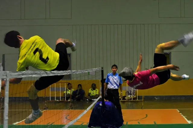 นักกีฬาตะกร้อสองฝั่งตาข่ายในโรงยิม คนหนึ่งสวมเสื้อเหลืองหมายเลข 2 กางเกงดำ อีกคนสวมเสื้อแดง กำลังกระโดดเตะลูกตะกร้อเหนือตาข่าย