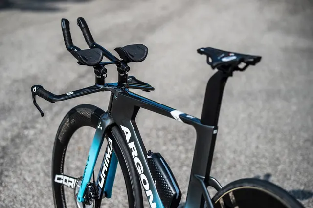จักรยานไทม์ไทรอัล Argon18 สีดำฟ้า เห็นแฮนด์ต่อ เบาะ ล้อคาร์บอนขอบสูงและกระติกน้ำใต้คาน วางบนพื้นยางมะตอย