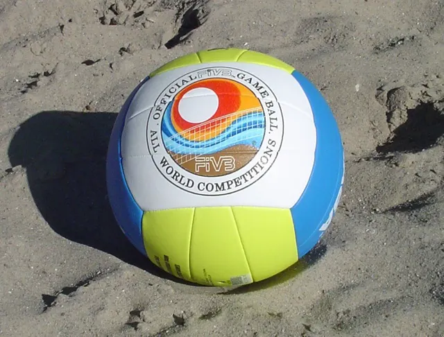 ลูกวอลเลย์บอลชายหาดสีขาว เหลืองและฟ้า วางบนพื้นทราย มีข้อความ Official FIVB Game Ball บนลูก